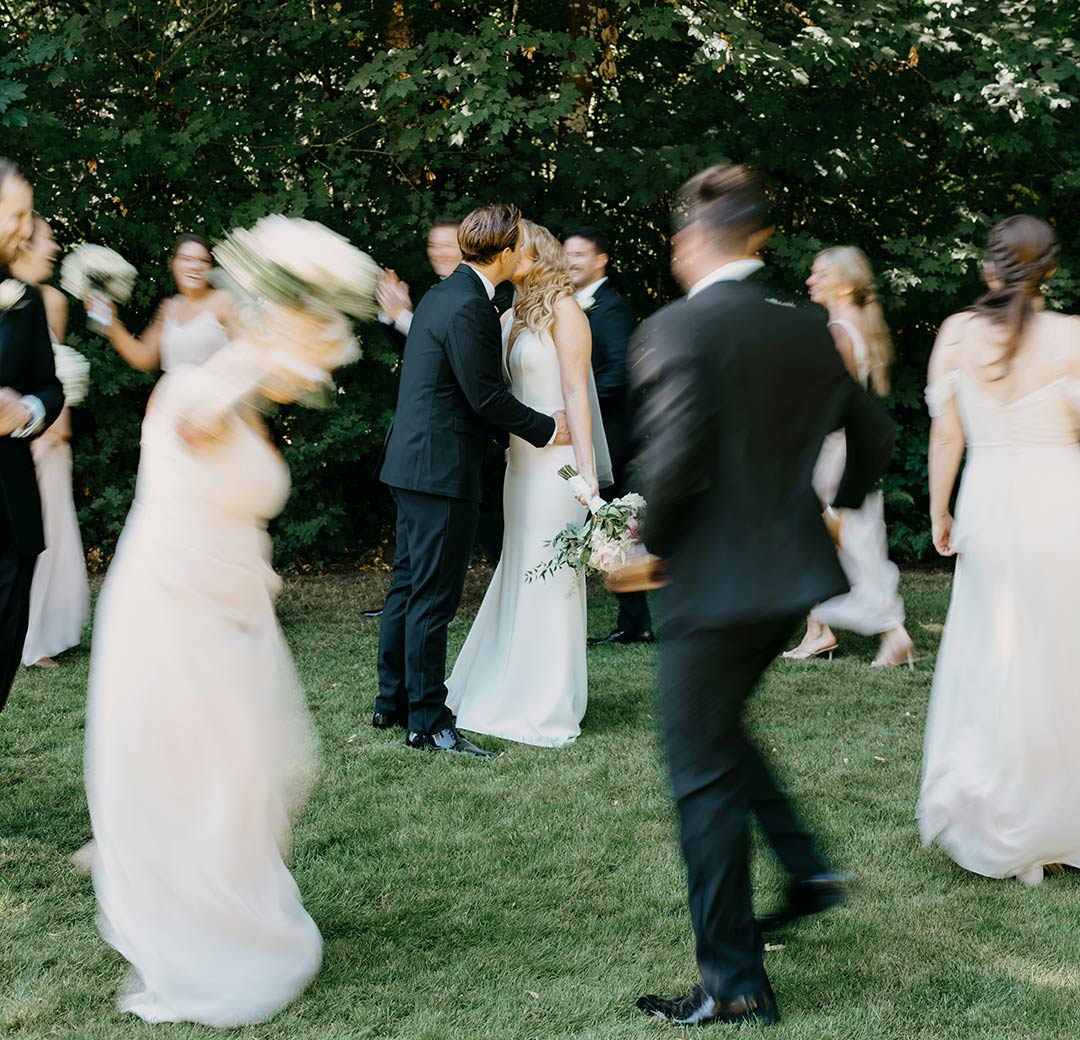 Bride and groom kiss on the lawn as their wedding party celebrates around them at an outdoor wedding near Stowe, VT